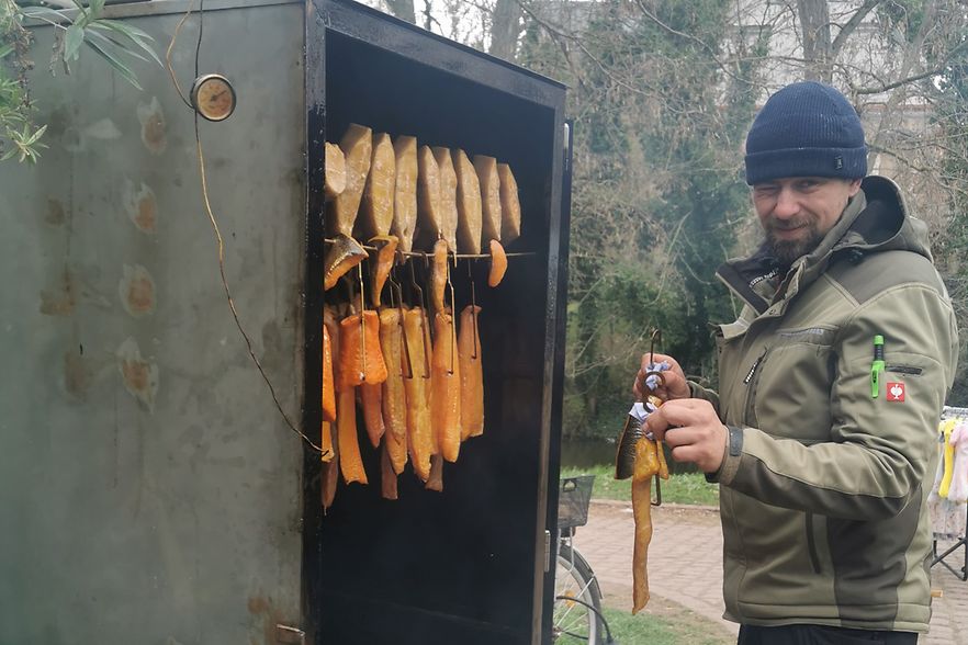 Ein Mann f&auml;delt eine Forelle auf einen Stab um ihn in den links zu sehenden R&auml;ucheroffen zu h&auml;ngen
