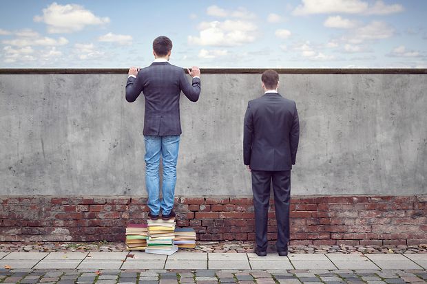 Zwei M&auml;nner vor Mauer, einer schaut mit Hilfe von B&uuml;chern dar&uuml;ber