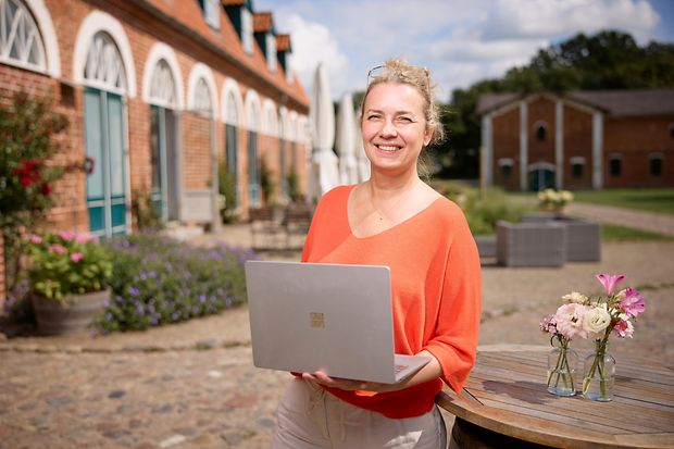 Person mit Laptop im Freien vor Geb&auml;ude, Blumen und Tischen