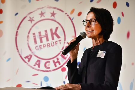 Eine Frau ist festlicher Kleidung steht auf einer B&uuml;hne und h&auml;lt eine Ansprache. 