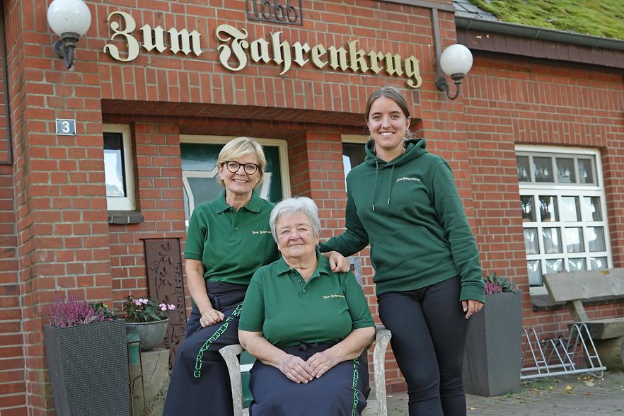 Drei Personen in gr&uuml;ner Arbeitskleidung vor dem Gasthaus &sbquo;Zum Fahrenkrug&lsquo;.