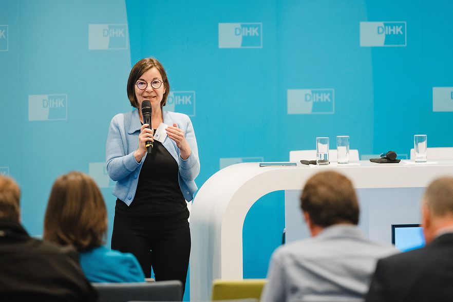 Eine Frau steht vor einem blauen Hintergrund. Sie h&auml;lt ein Mikrofon in der Hand und spricht zu einem Publikum.