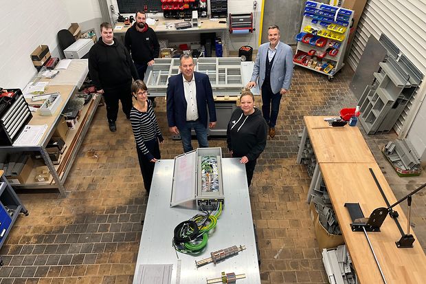 Aufnahme von oben in einen Produktionsraum mit Metalltischen und elektronischen ger&auml;ten. F&uuml;nf Menschen schauen nach oben
