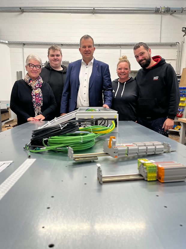 Fünf Menschen stehen in einer Reihe hinter einem Metalltisch. Auf dem Tisch liegen elektronische Geräte.