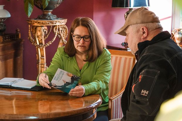 Eine Frau und ein Mann sitzen an einem Tisch. Die Frau erkl&auml;rt dem Mann die Inhalte eines Flyers.