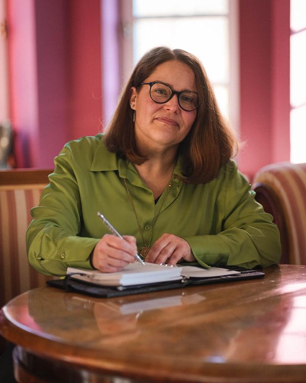 Eine Frau sitzt an einem Tisch. Sie hat einen Stift in der Hand und vor sich eine Dokumentenmappe.