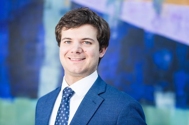 Ein Mann mit blauem Anzug steht vor einer bunten Wand und lächelt in die Kamera.