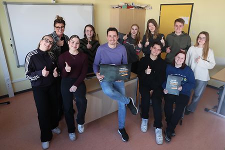 Schulklasse posiert gemeinsam in einem Klassenraum und zeigt Daumen nach oben, einige halten Mappen eines Bildungsprojekts.