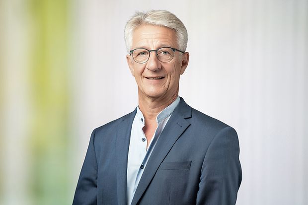 Ein Mann im grauen Anzug steht vor einem graugr&uuml;nen Hintergrund und schaut in die Kamera.