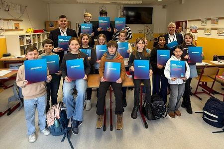 Sch&uuml;lerinnen und Sch&uuml;ler sind in einem Klassenraum und halten Zeugnismappen in den H&auml;nden. Zwei M&auml;nner stehen dabei.