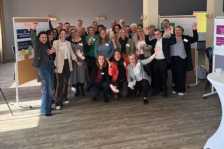 Gruppe von Workshop-Teilnehmenden steht gut gelaunt in einem hellen Raum vor Moderationstafeln und winkt in die Kamera.