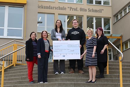 Gruppe h&auml;lt einen gro&szlig;en Spendenscheck vor der Sekundarschule &bdquo;Prof. Otto Schmeil&ldquo;.