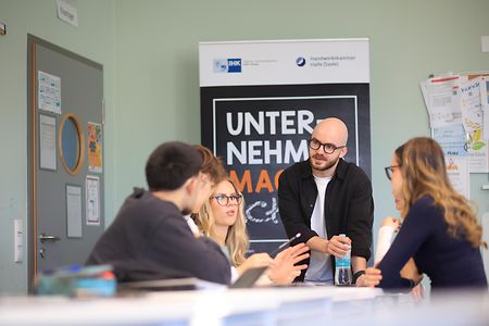 Personengruppe im Klassenraum diskutiert vor einem Roll‑Up der Initiative Unternehmer machen Schule.