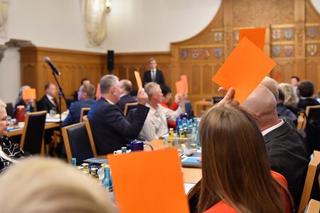 Personen in einem Sitzungssaal heben orange Karten zur Abstimmung.