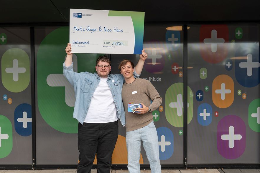 Zwei M&auml;nner halten einen Preisgeldscheck hoch und l&auml;cheln in die Kamera.
