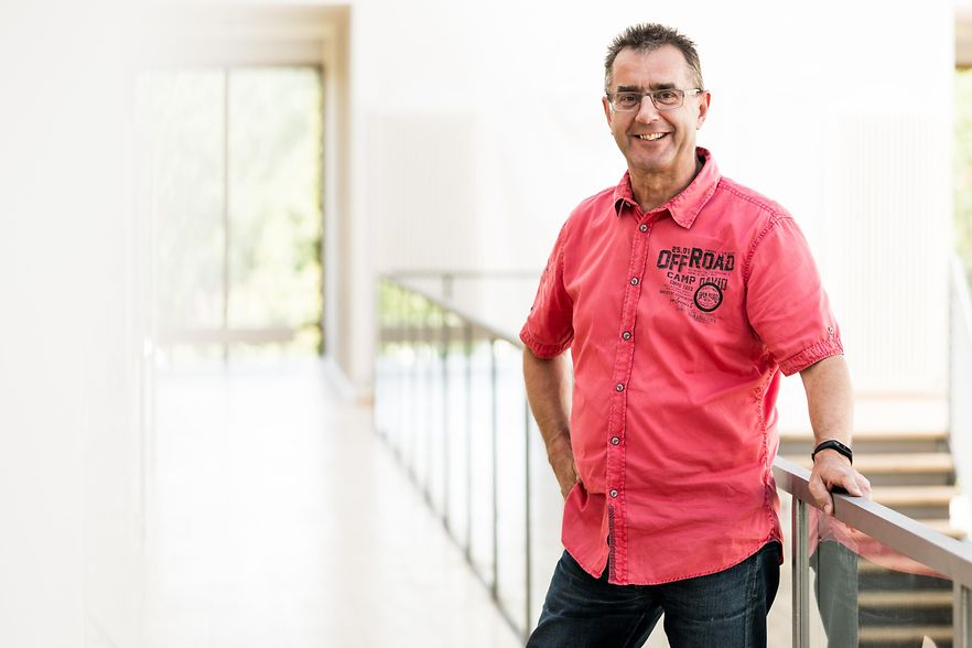 Ein Mann in rotem Shirt lehnt sich an ein Gel&auml;nder und l&auml;chelt in die Kamera.