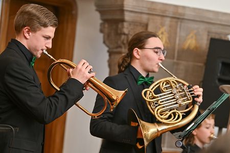 Zwei Musiker in schwarzen Anz&uuml;gen spielen Trompete und Horn bei einem Konzert in einem Saal.