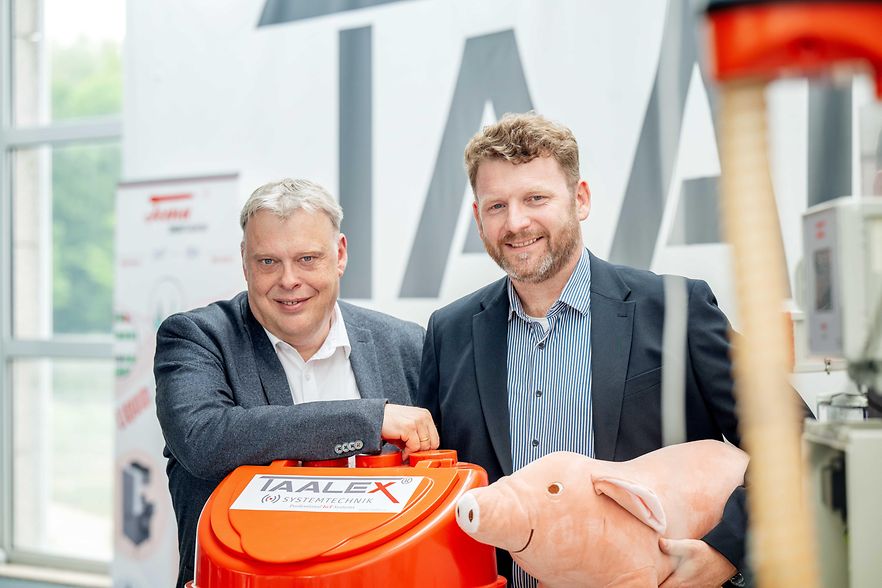Zwei M&auml;nner hinter einer Maschine f&uuml;r Landwirtschaft, mit einem Pl&uuml;schschwein.