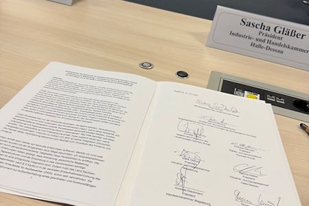 Offenes Dokument mit Unterschriften auf einem Konferenztisch, dahinter Namensschilder und Personen in einem Besprechungsraum.