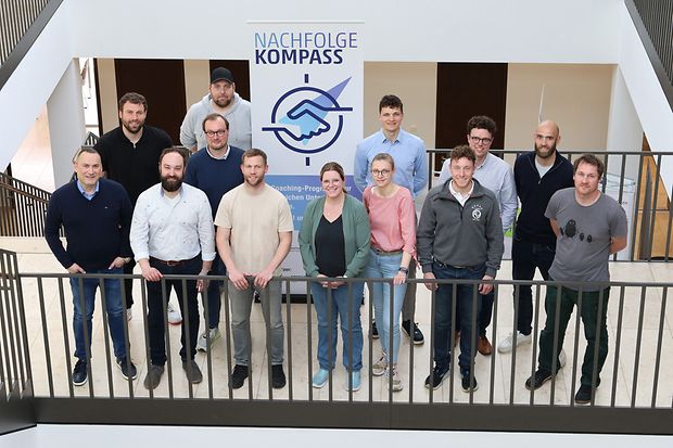 13 Menschen stehen vor einem Aufsteller mit der Aufschrift "NachfolgeKompass"