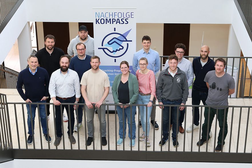 13 Menschen stehen vor einem Aufsteller mit der Aufschrift "NachfolgeKompass"