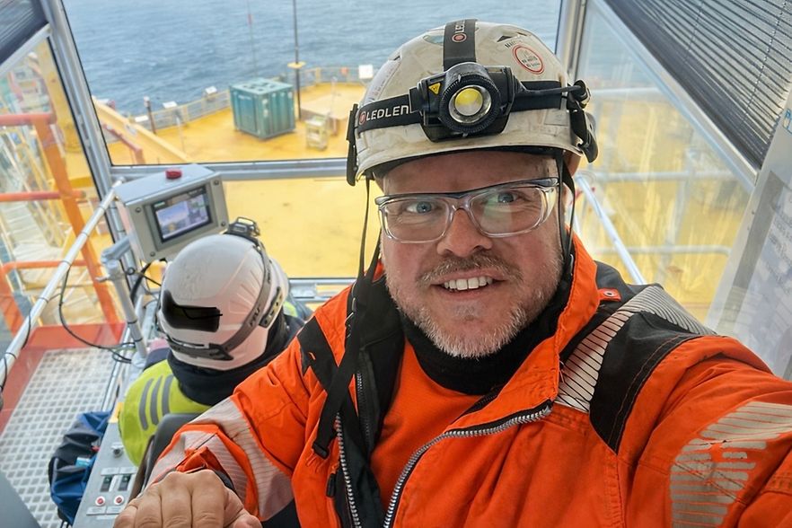 Ein Mann mit Arbeitskleidung und Helm ist im Vordergrund. Im Hintergrund eine Offshore-Plattform und das Meer.