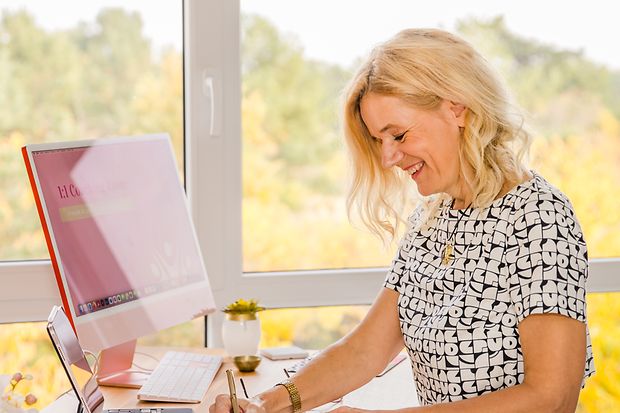 Eine blonde Frau sitzt vor einem Computer. Vor sich hat sie ein Notizbuch und ein Handy.