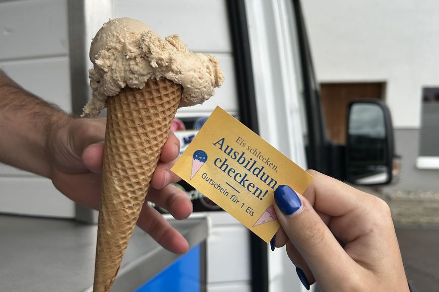 Ein Eisverk&auml;ufer reicht eine Kugel Eis in der Waffel an eine Person. Die Person reicht einen Gutschein.