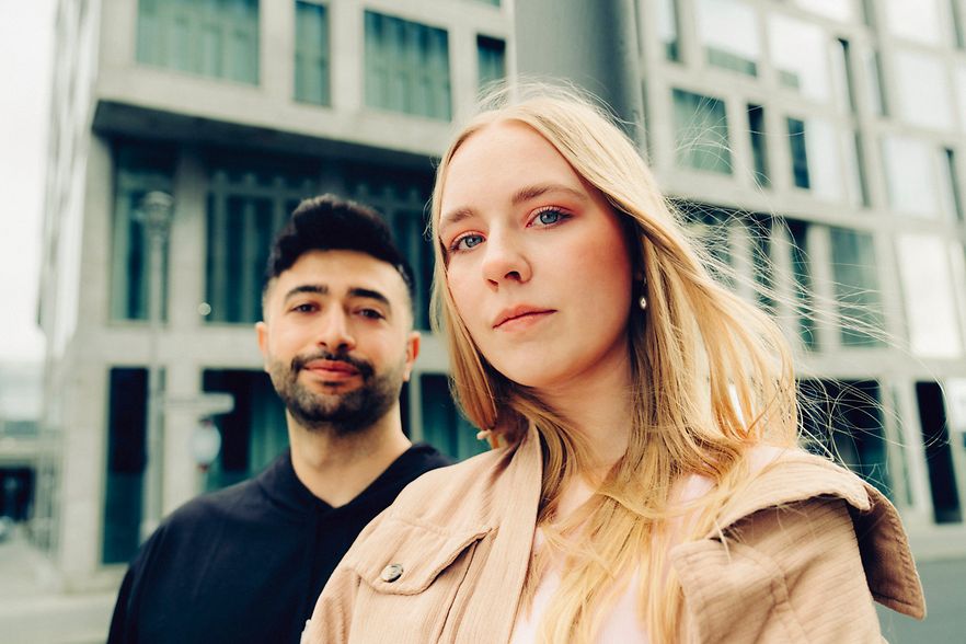 Ein junger Mann steht schr&auml;g hinter einer jungen, blonden Frau.