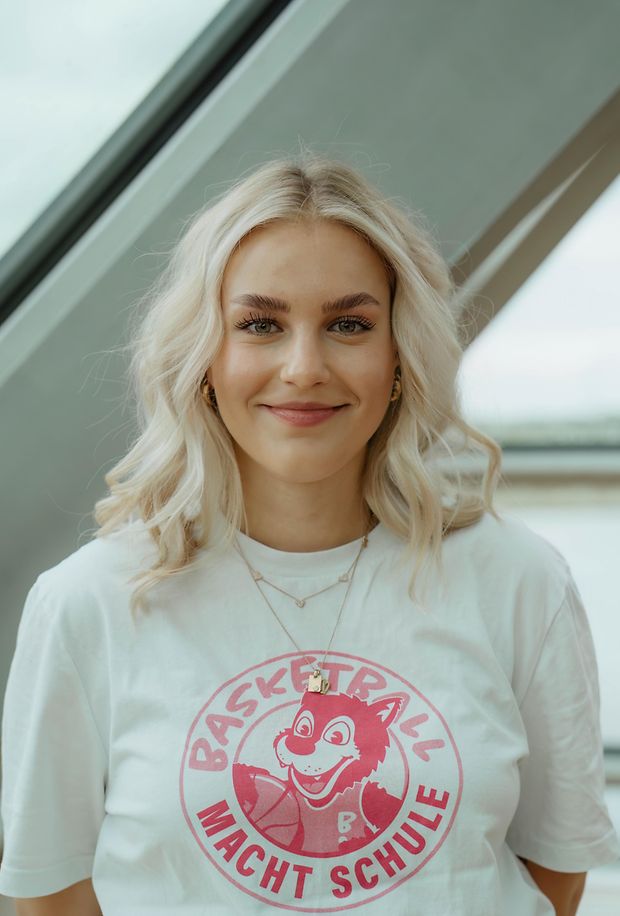 Eine junge Frau mit blonden Haaren lächelt in die Kamera. Sie trägt ein T-Shirt mit der Aufschrift Basketball macht Schule.